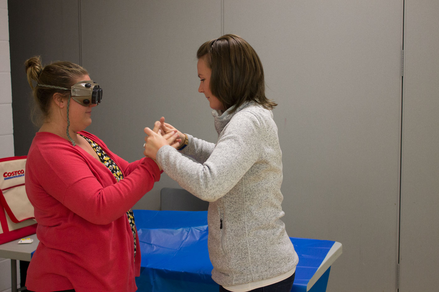 Two women stand across from one another, acting out a simulation exercise. One woman wears simulation goggles as they communicate with hand-over-hand sign language.