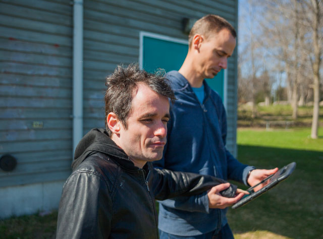 An intervenor and man with deafblindness stand outside on a sunny day. The intervenor assists the man with communicating through an iPad.