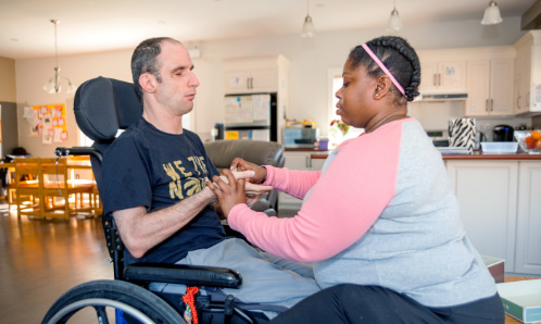 Un homme est assis devant son interprète tactile. L’homme est en fauteuil roulant. Les deux communiquent par le toucher.