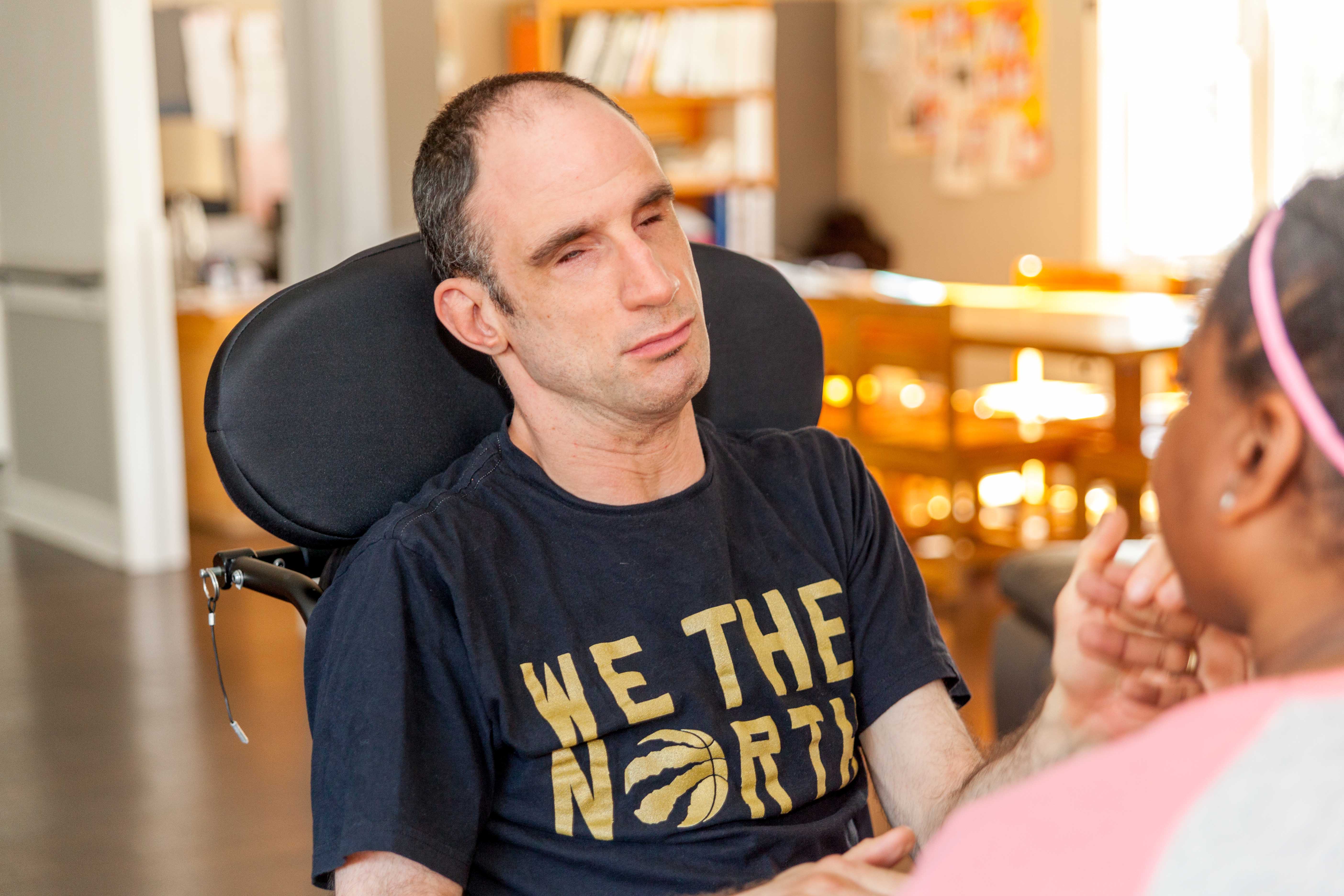 Un homme atteint de surdicécité est assis dans son fauteuil roulant et communique par le toucher avec son interprète tactile qui est en grande partie hors champ.