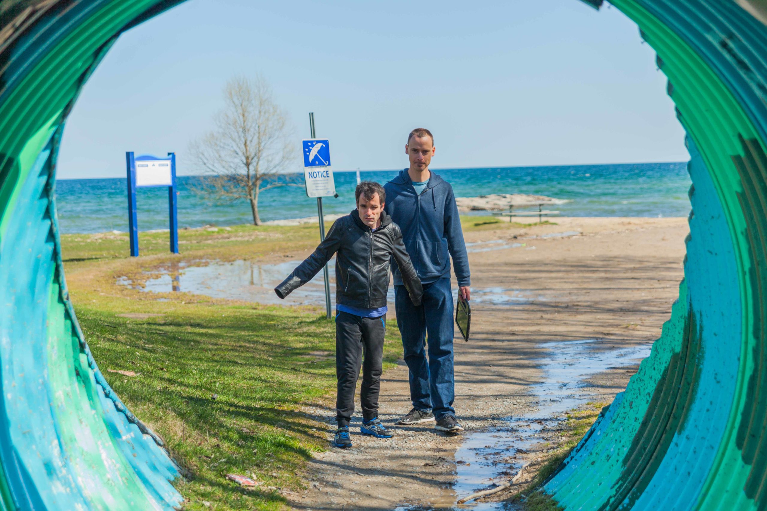 This image is captured through the perspective of a turquoise tunnel. An intervenor guides a man with deafblindness as they walk on a sandy beach.