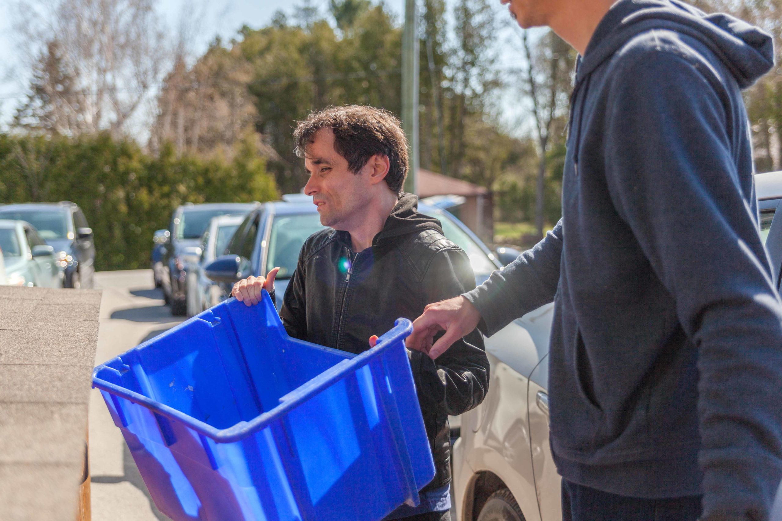 Intervenor assists man with deafblindness in taking out the recycling. They each hold an end of the blue recycle bin.
