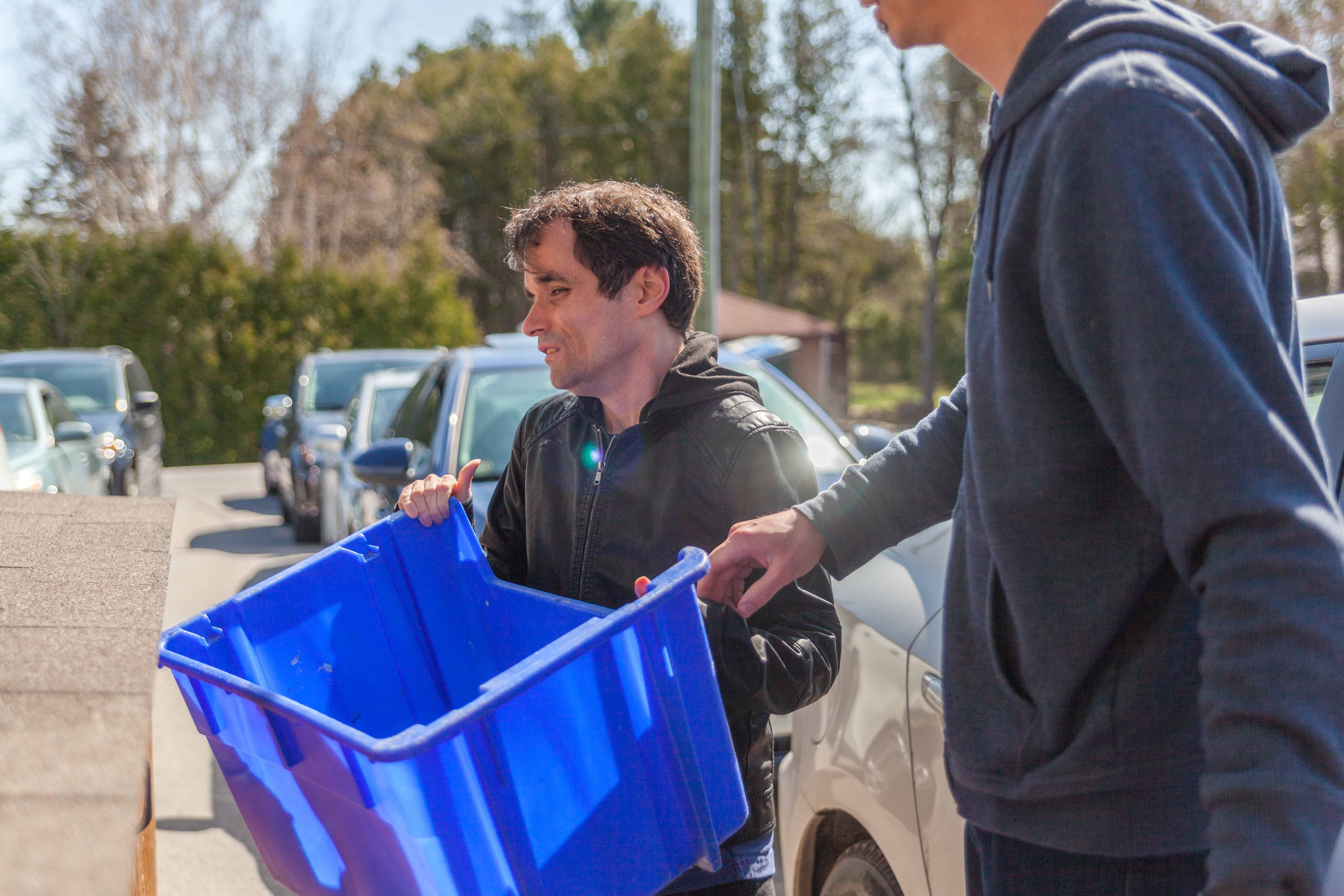 Un homme atteint de surdicécité sort le recyclage avec l’aide de son interprète tactile. Chaque personne tient un bout du bac bleu.