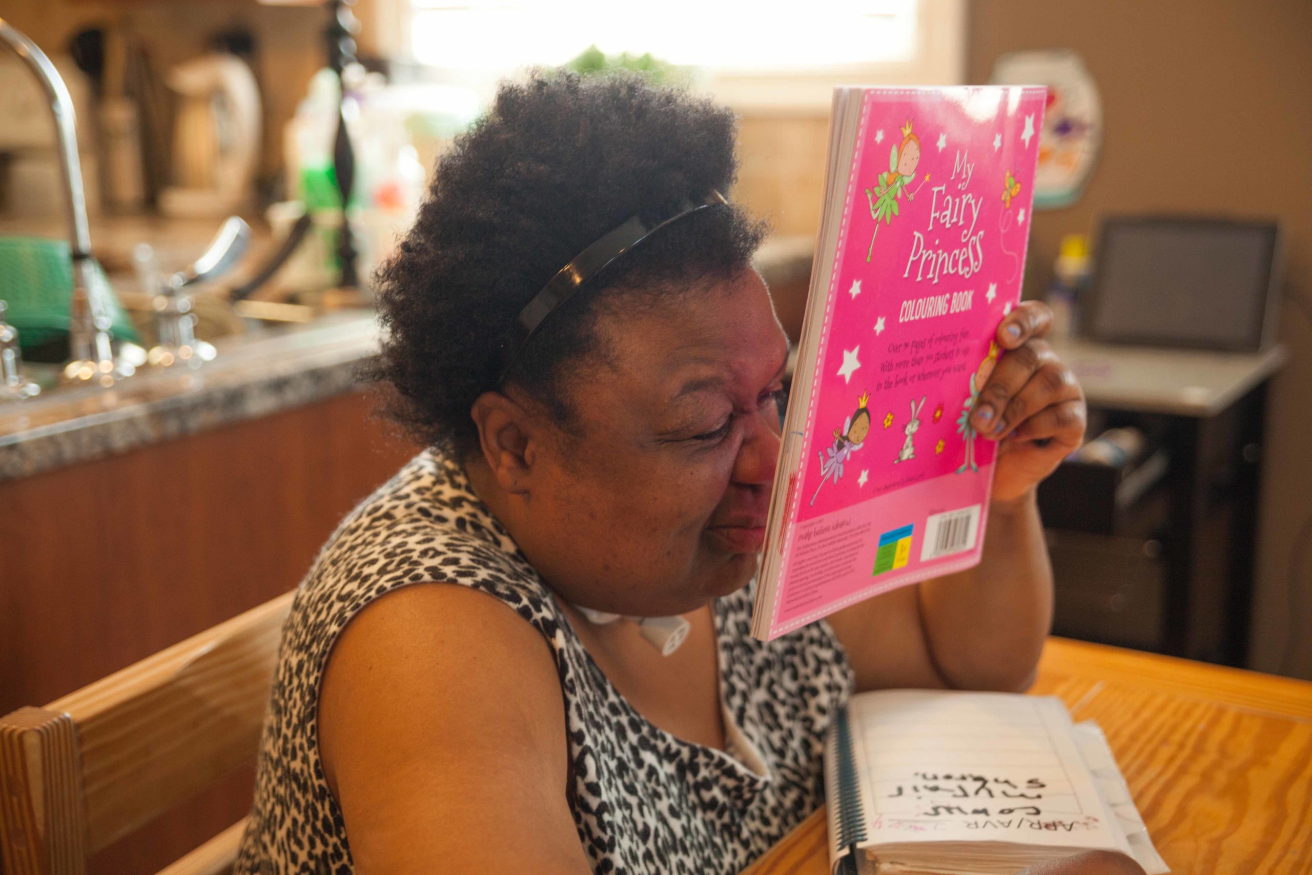 A woman with deafblindness sits at a kitchen table. She holds a pink book very close to her eye so that she can read it.