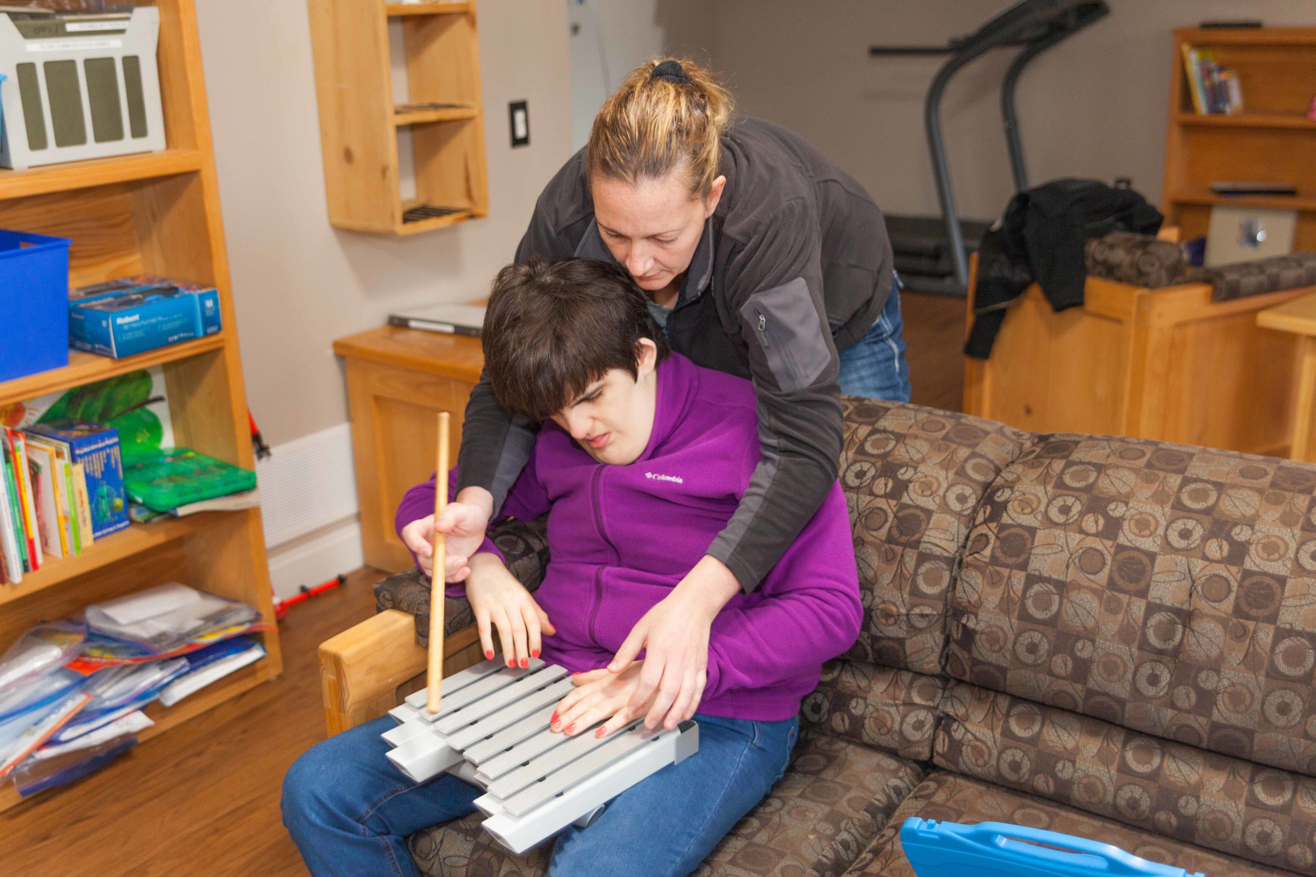 Intervenor stands behind a woman with deafblindness. The intervenor holds the woman’s hands, supporting her in playing the xylophone.