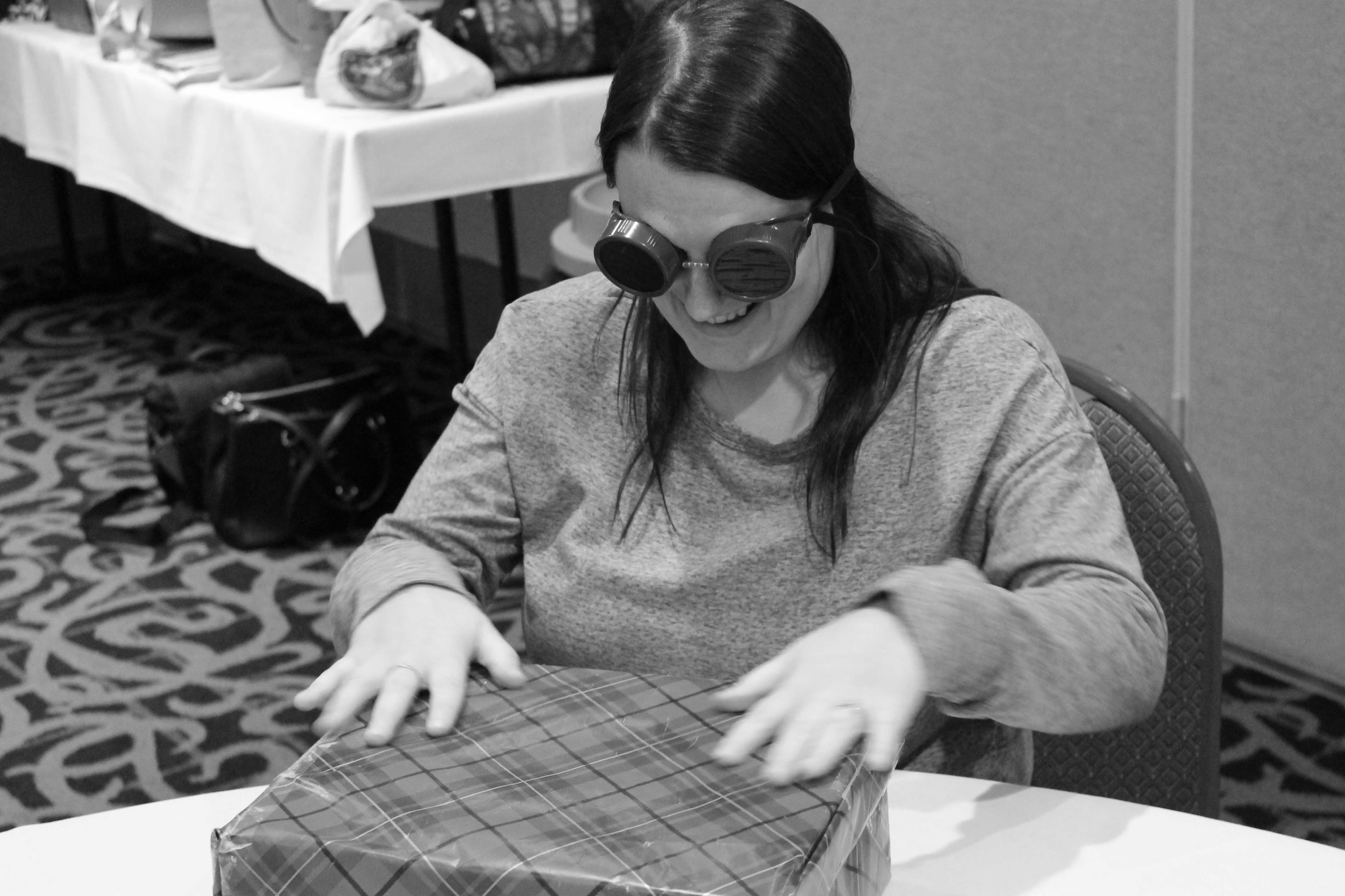 Une photo en noir et blanc d’une jeune femme assise à une table durant un exercice de formation par simulation. Elle porte des lunettes de simulation et manipule un « cadeau » carré, emballé dans du papier à motifs.