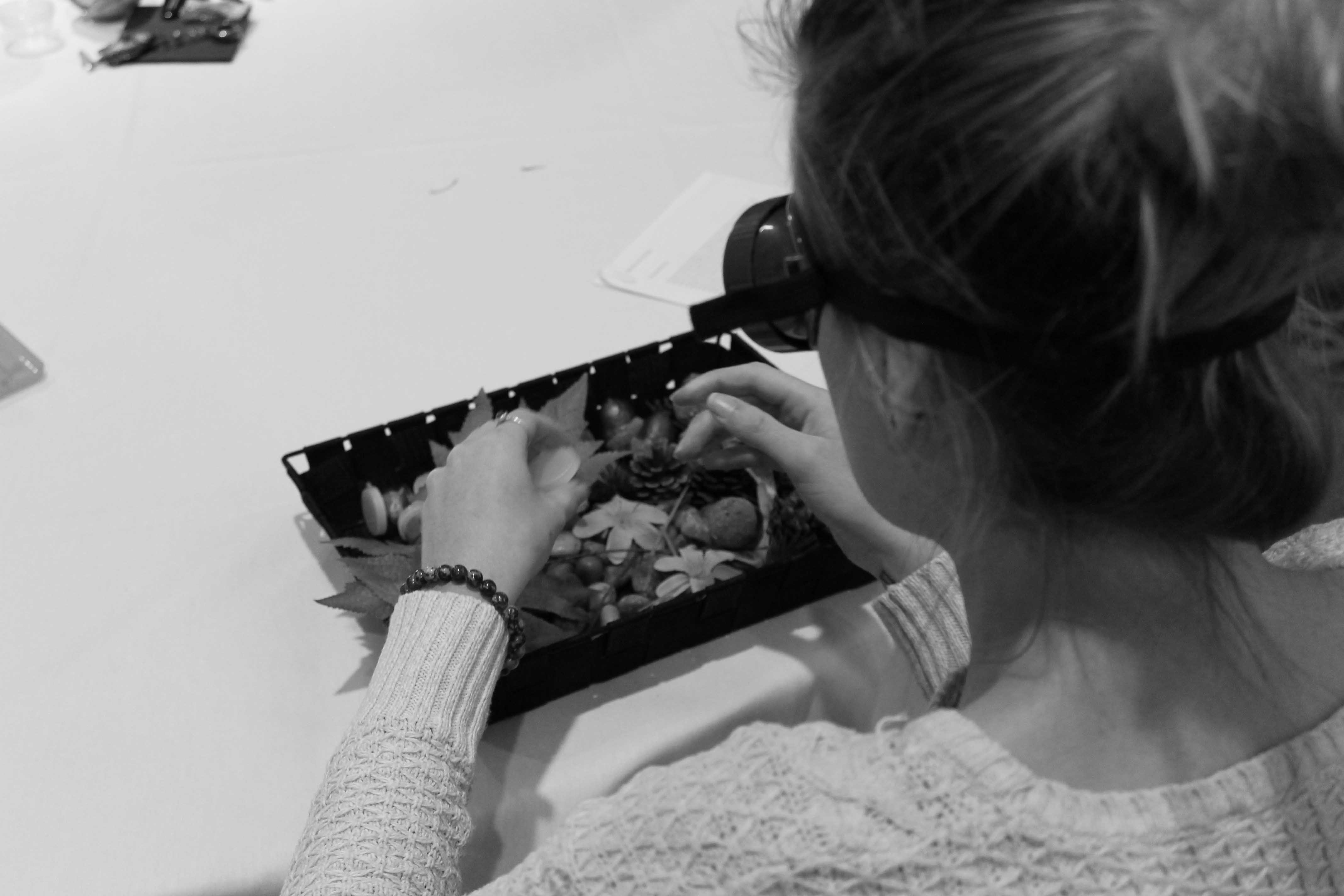 Image en noir et blanc prise par-dessus l’épaule d’une femme effectuant un exercice de formation par simulation. Elle porte des lunettes de simulation et manipule des objets sensoriels dans une boîte.