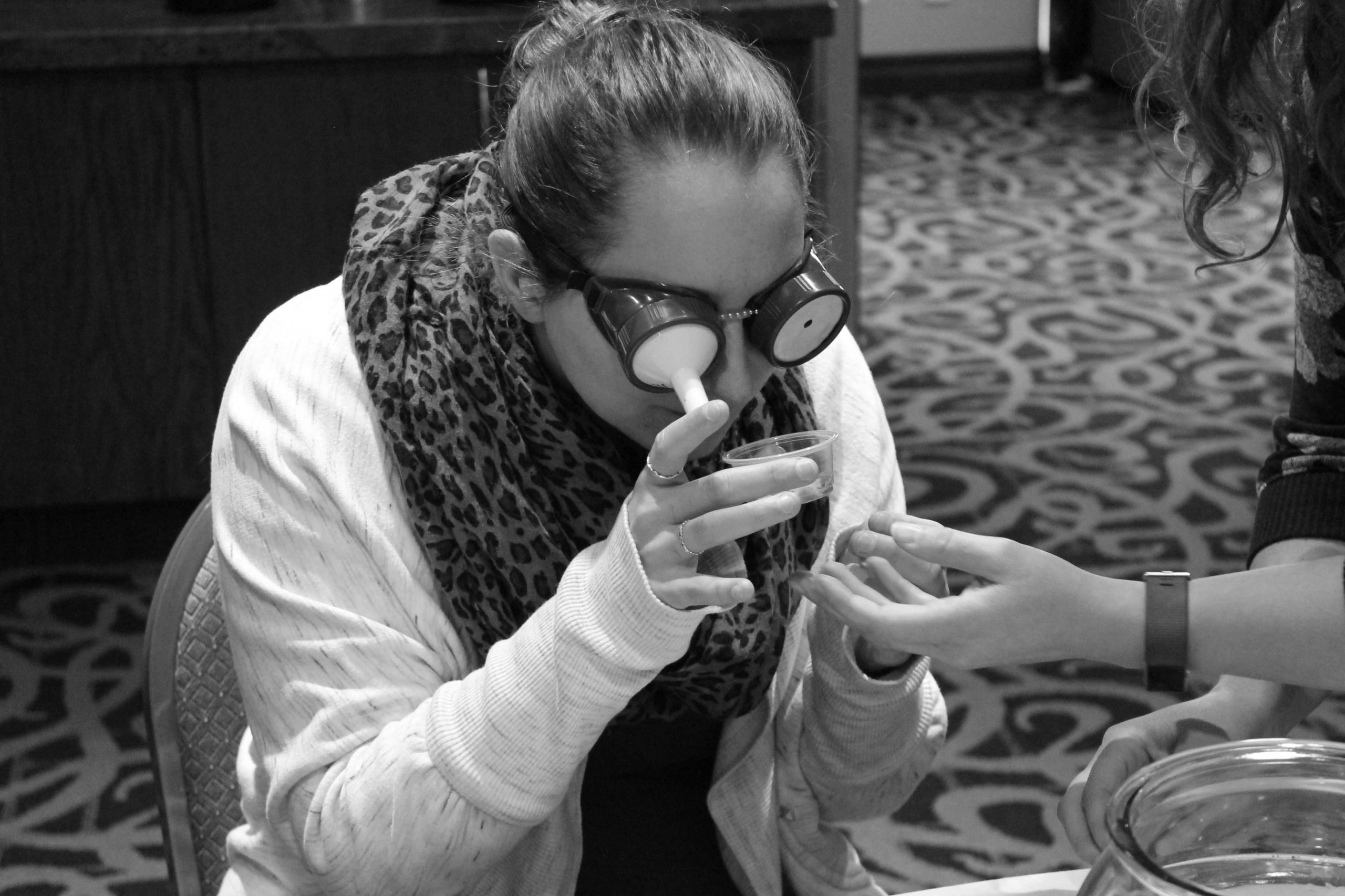 A black and white photo of a young woman participating in a simulation training exercise. She is wearing simulation goggles while examining an object. The goggles help her to understand the functional impact of visual impairment or low vision.