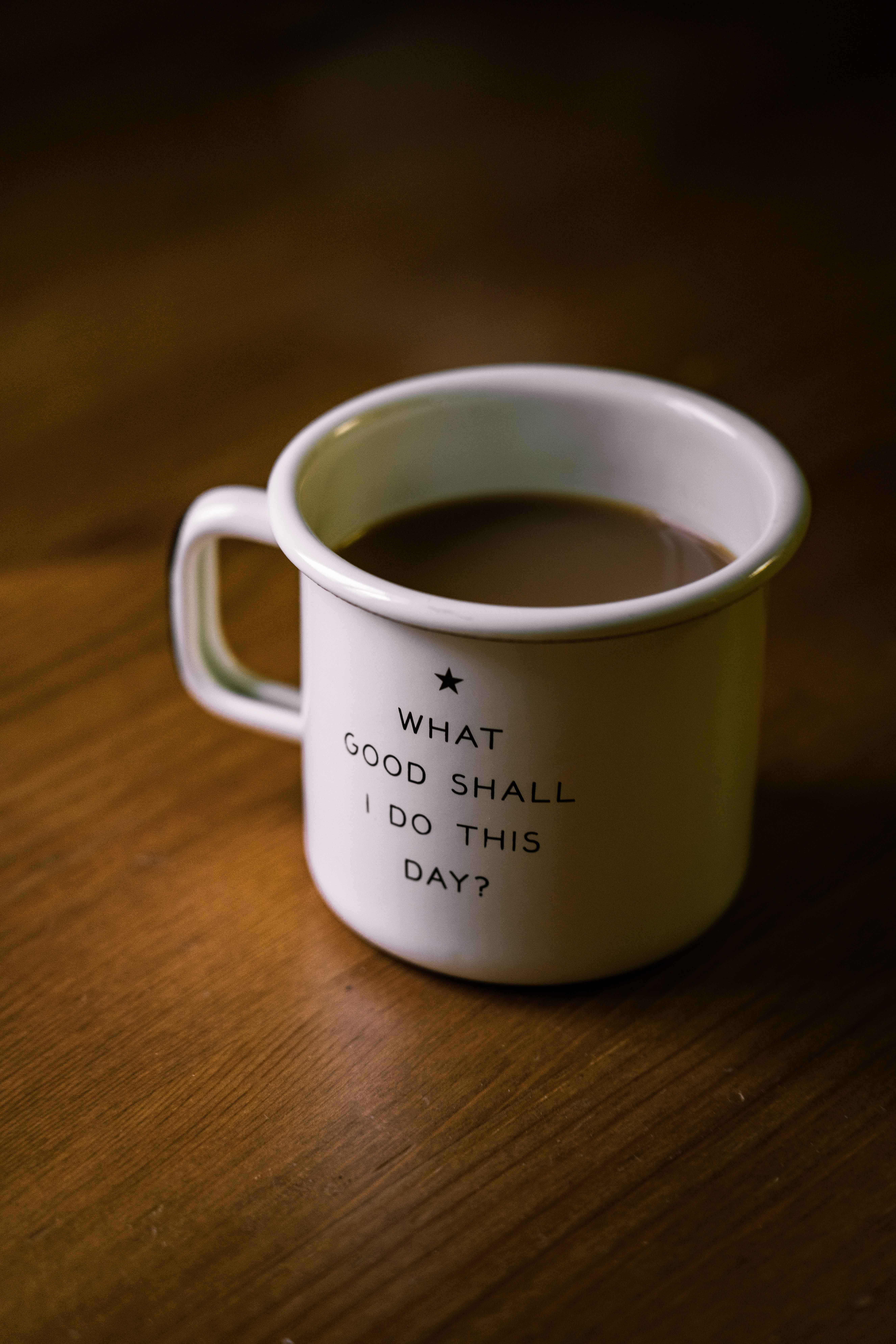 Mug with coffee in it on a table. The mug has decorative text that says "What good shall I do this day?"