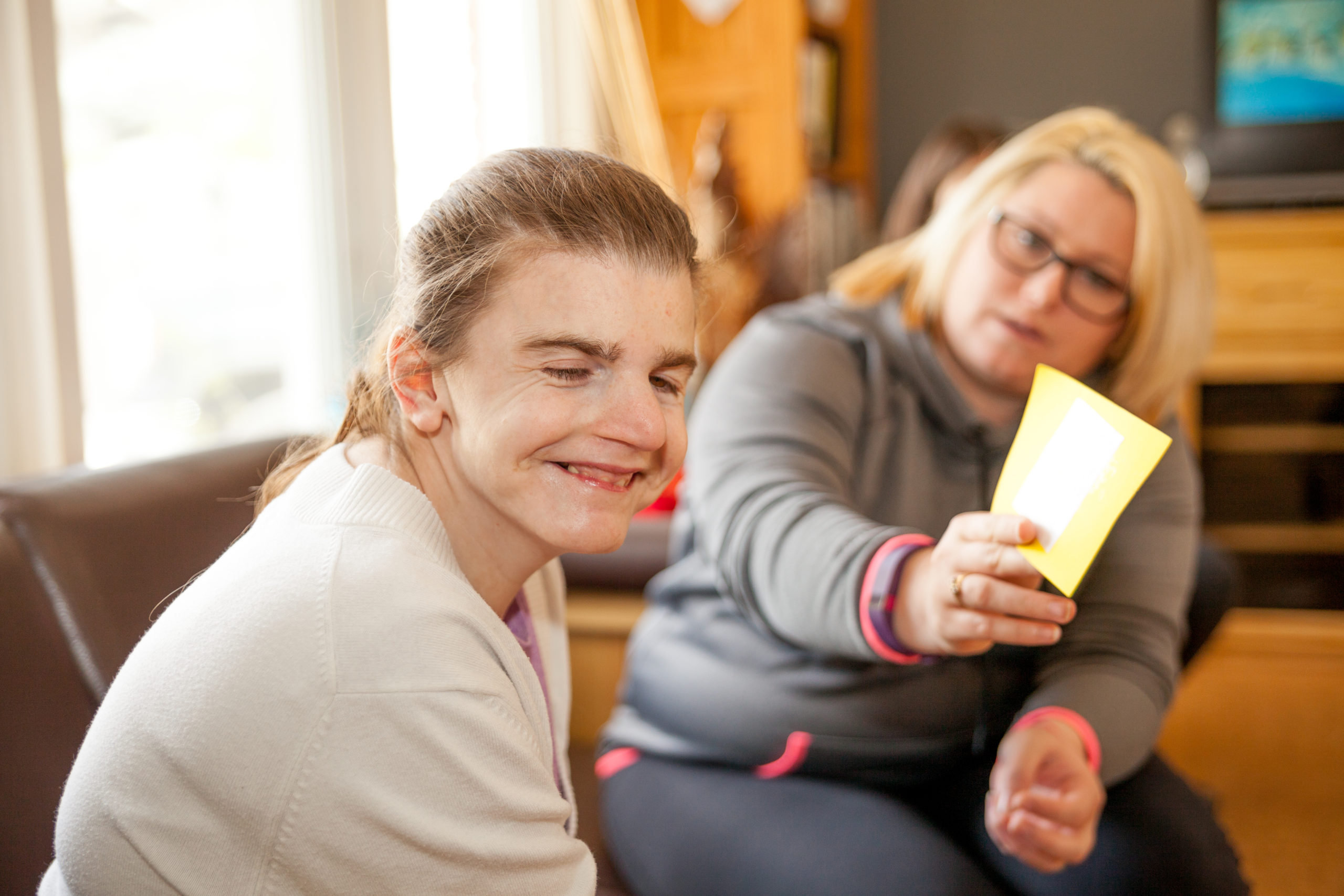 Intervenor sits with person supported on a couch, looking at a booklet. Person supported smiles at the camera.