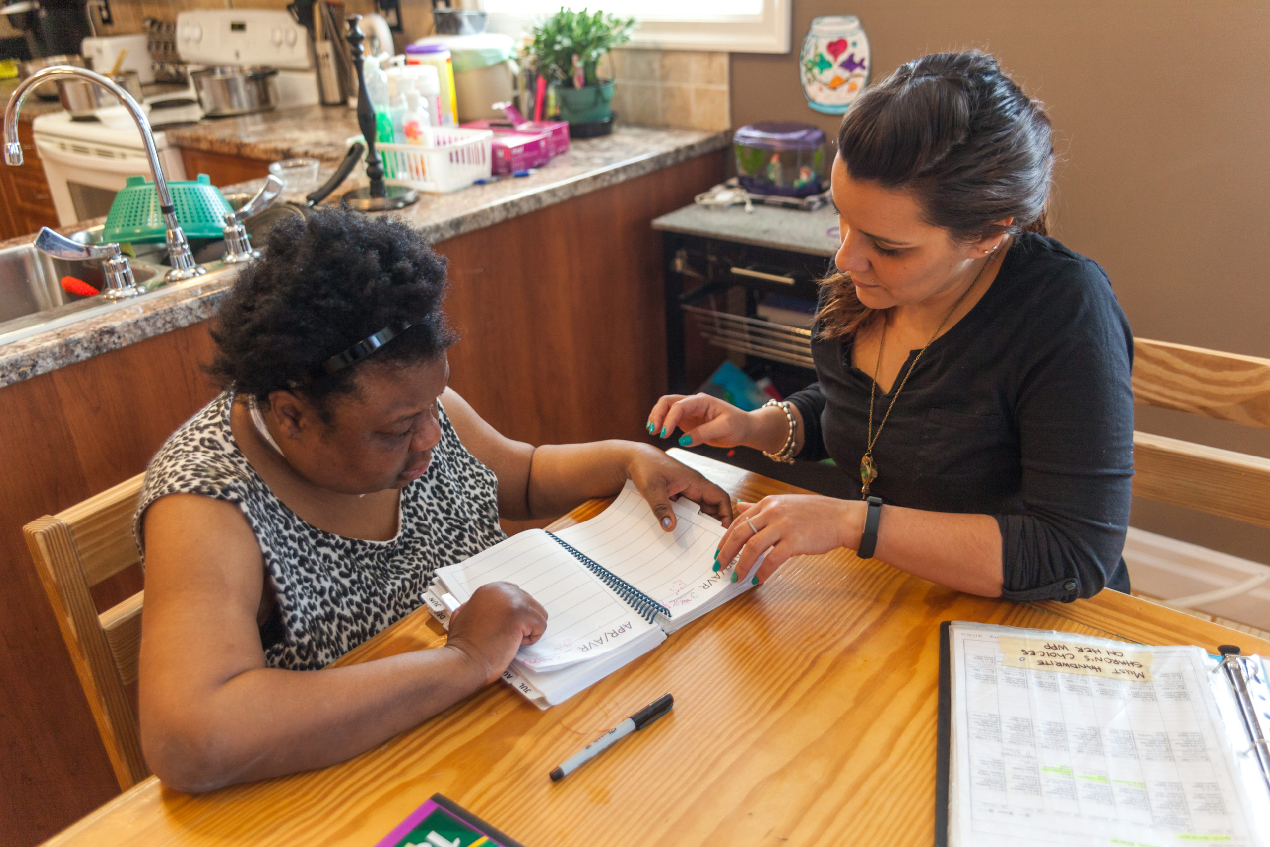 Intervenor sits with the woman she is supporting at a kitchen table, the two interact as the woman writes in a notebook.