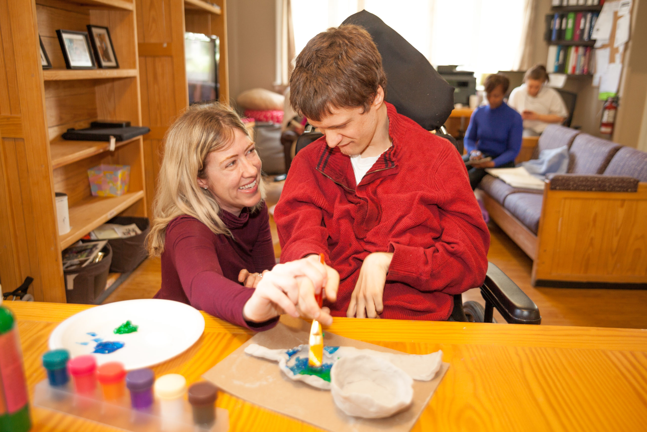 Intervenor smiling assisting a man in painting a craft at the kitchen table.