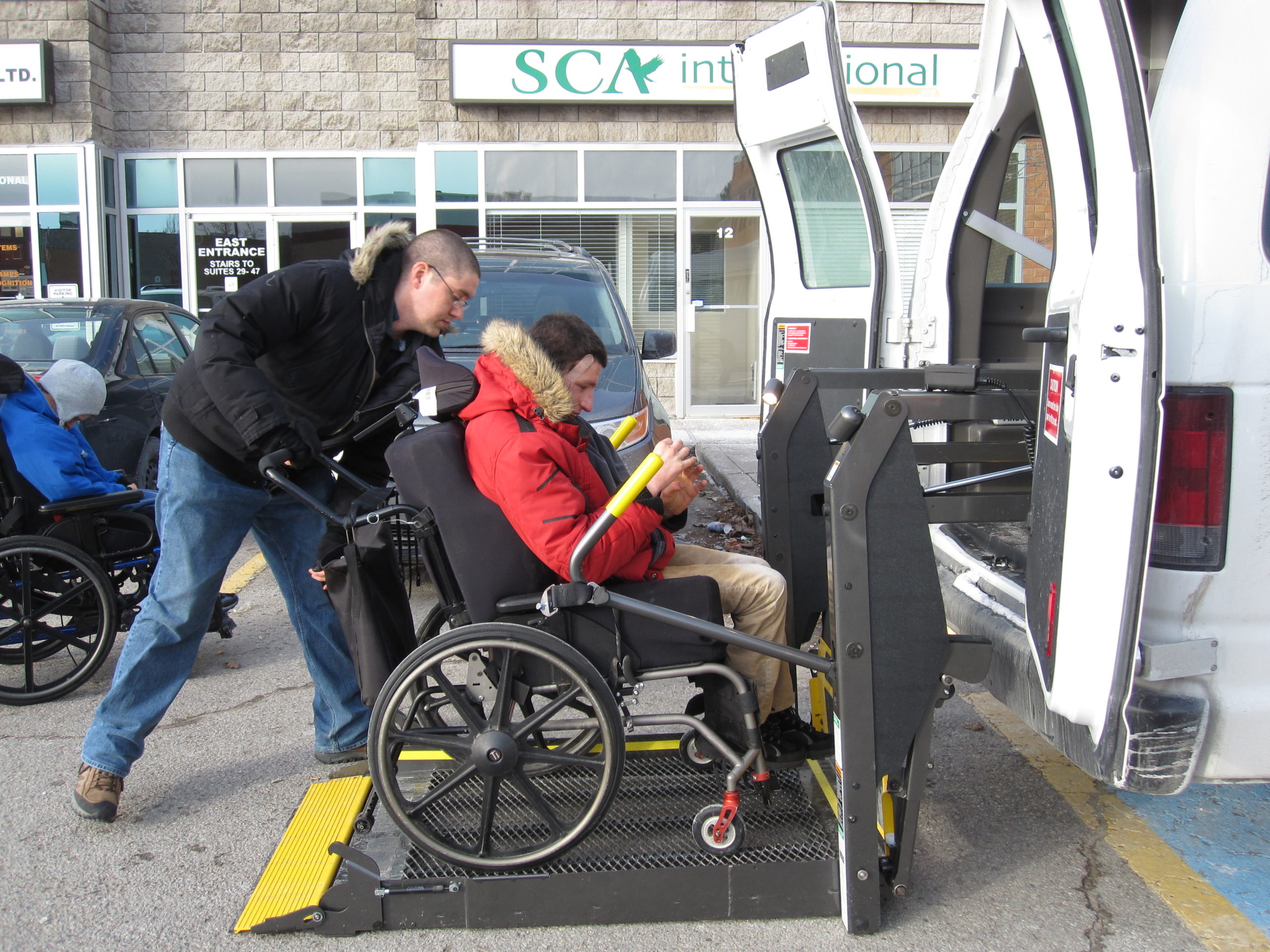 L’interprète tactile pousse un jeune homme en fauteuil roulant sur une rampe d’accès à une camionnette accessible.