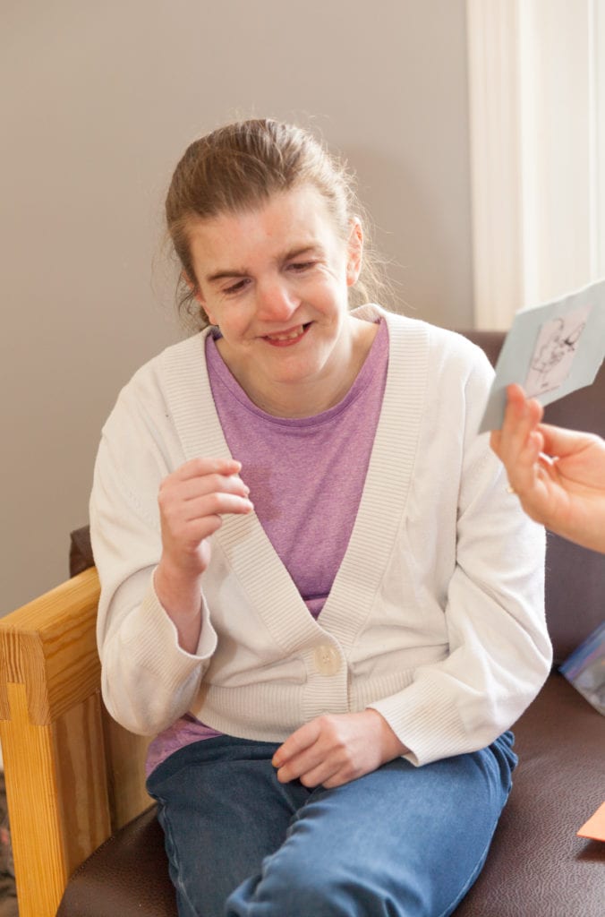 A young woman, sits on a couch smiling with her right arm raised to chest level. She is looking at a paper held by an intervenor not photographed.