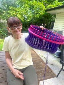 A woman holds up a round knitting loom with knitted purple yarn