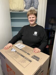 Sara is shown preparing to pack some of her things for her move. She is smiling and standing in front of a linen closet with a cardboard box.